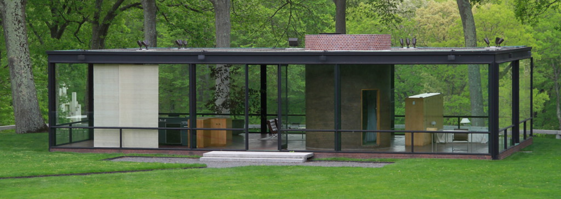 Glass House, New Canaan, CT. Black steel framed modern home with transparent walls, on a green lawn with trees in the background.
