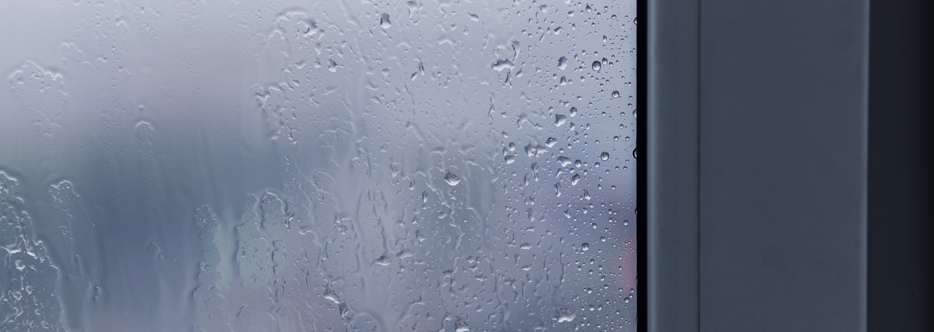 Raindrops on a window with a blurry outdoor background and a grey frame.