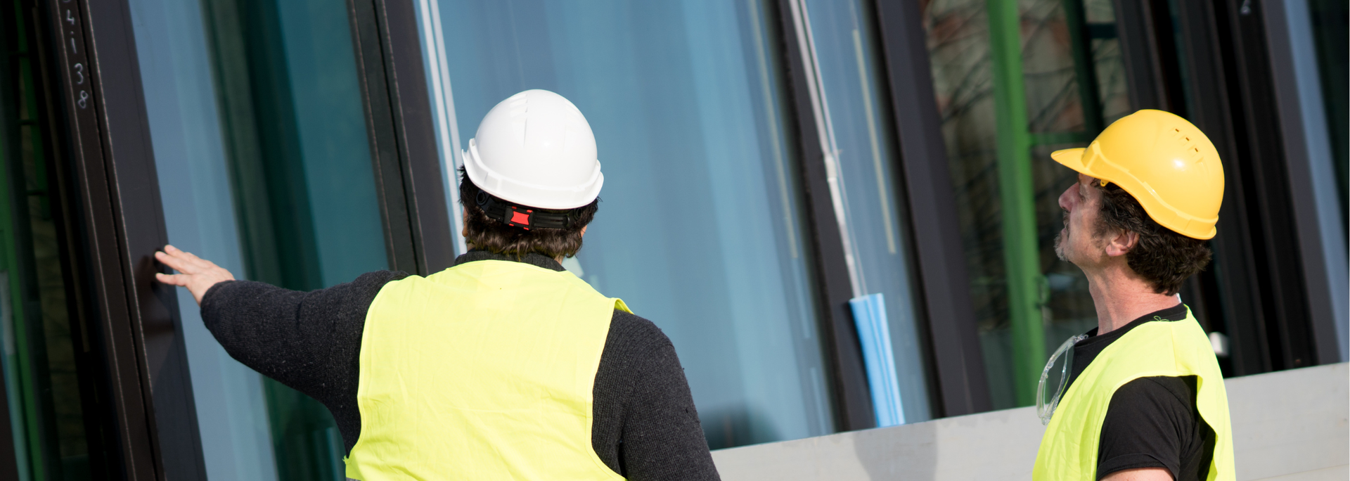 Two construction workers, one in white hard hat and other in yellow, examining glass panels.