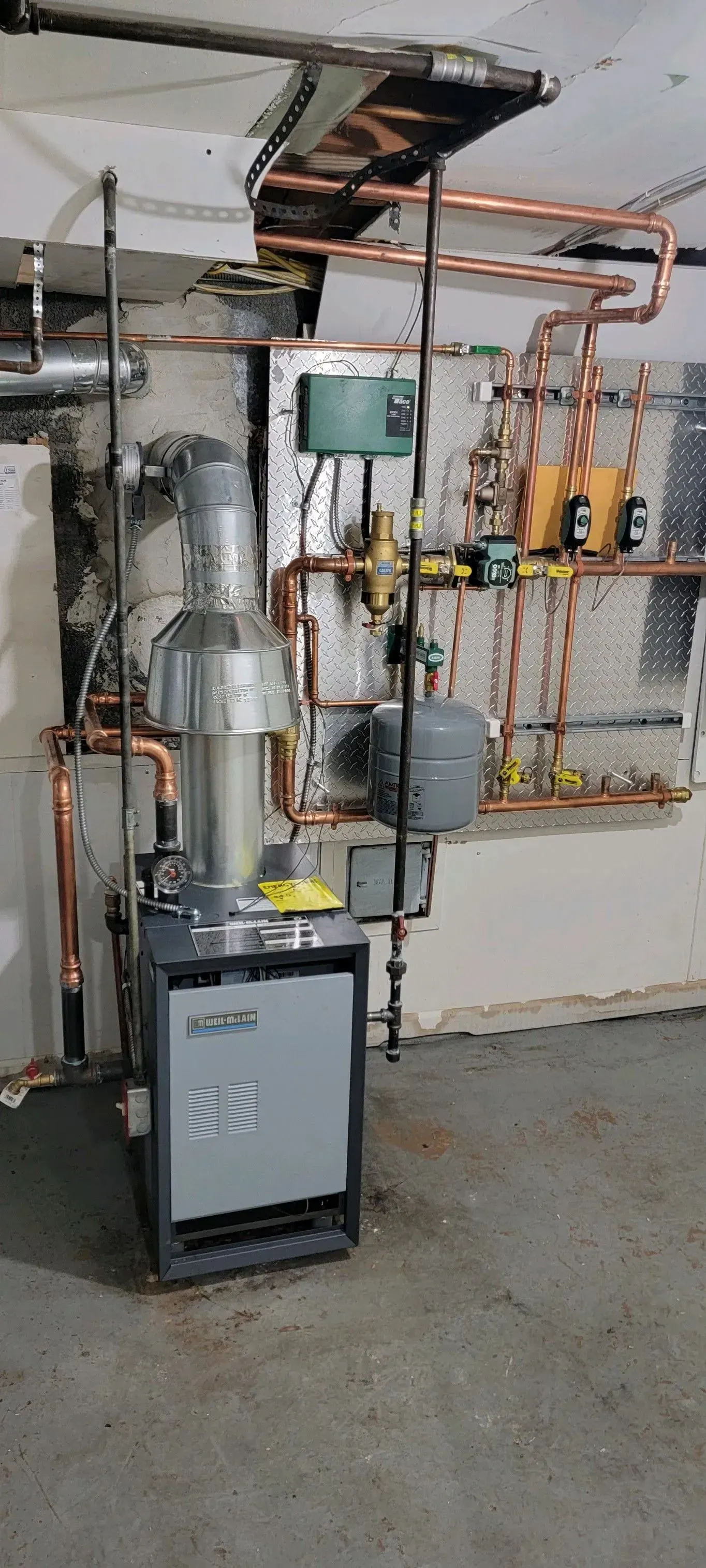 A boiler is sitting in a basement next to a wall with copper pipes.