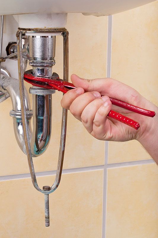 Person using pliers to work on a sink's plumbing. Red pliers are used on a metal pipe under a white sink.