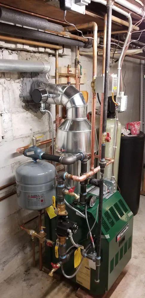 A green boiler is sitting in a basement surrounded by pipes.