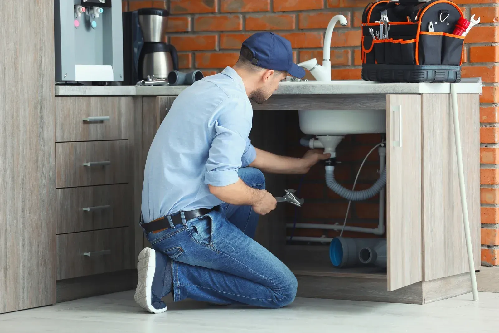 Plumber in blue shirt and jeans repairs pipes under kitchen sink near tools and brick wall.