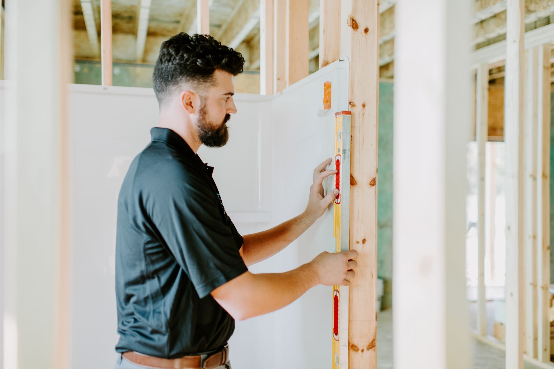 Man in black shirt uses a level on wood framing in a construction site, preparing to install drywall.