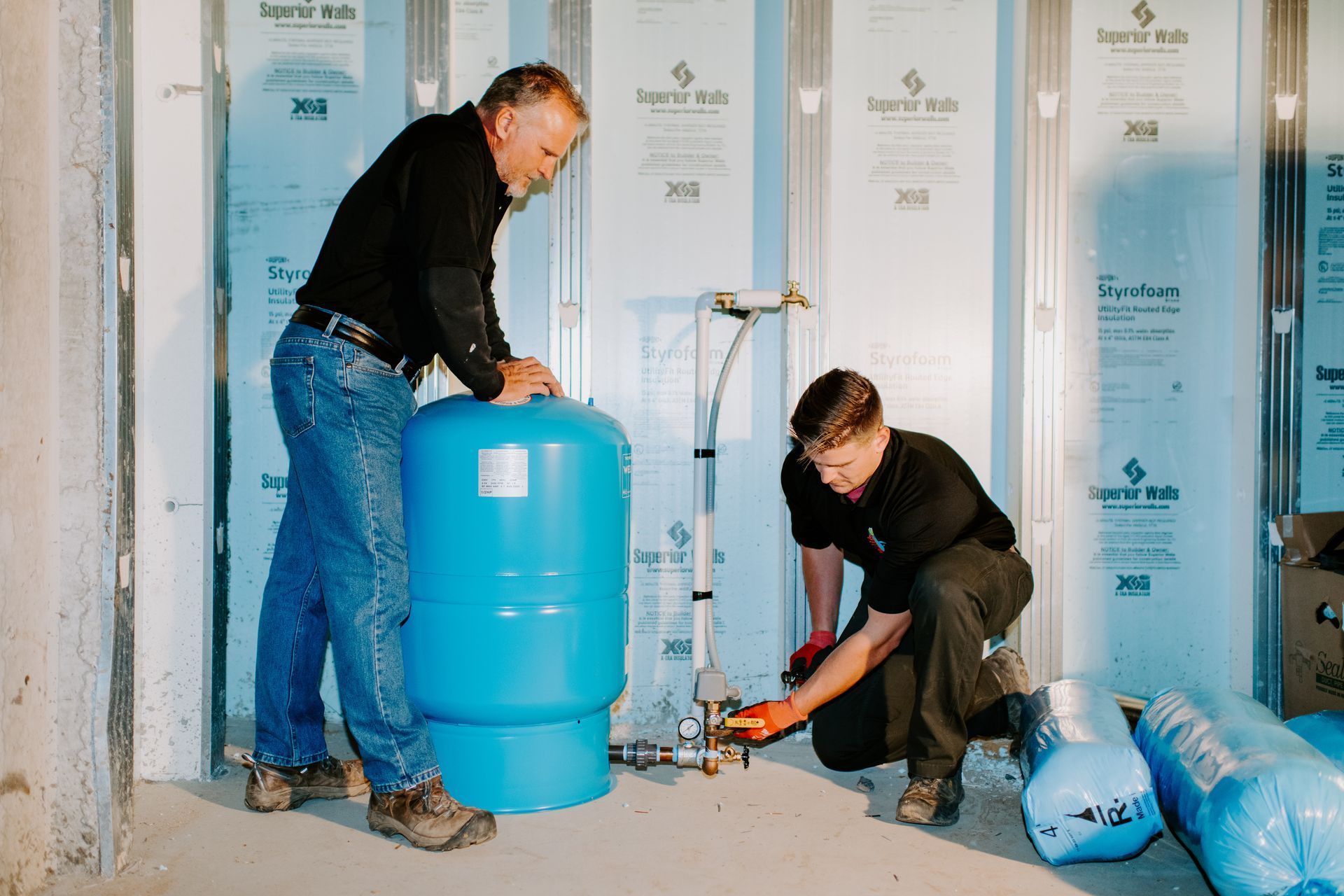 Two men installing plumbing in a basement; blue tank, pipes, and insulation.