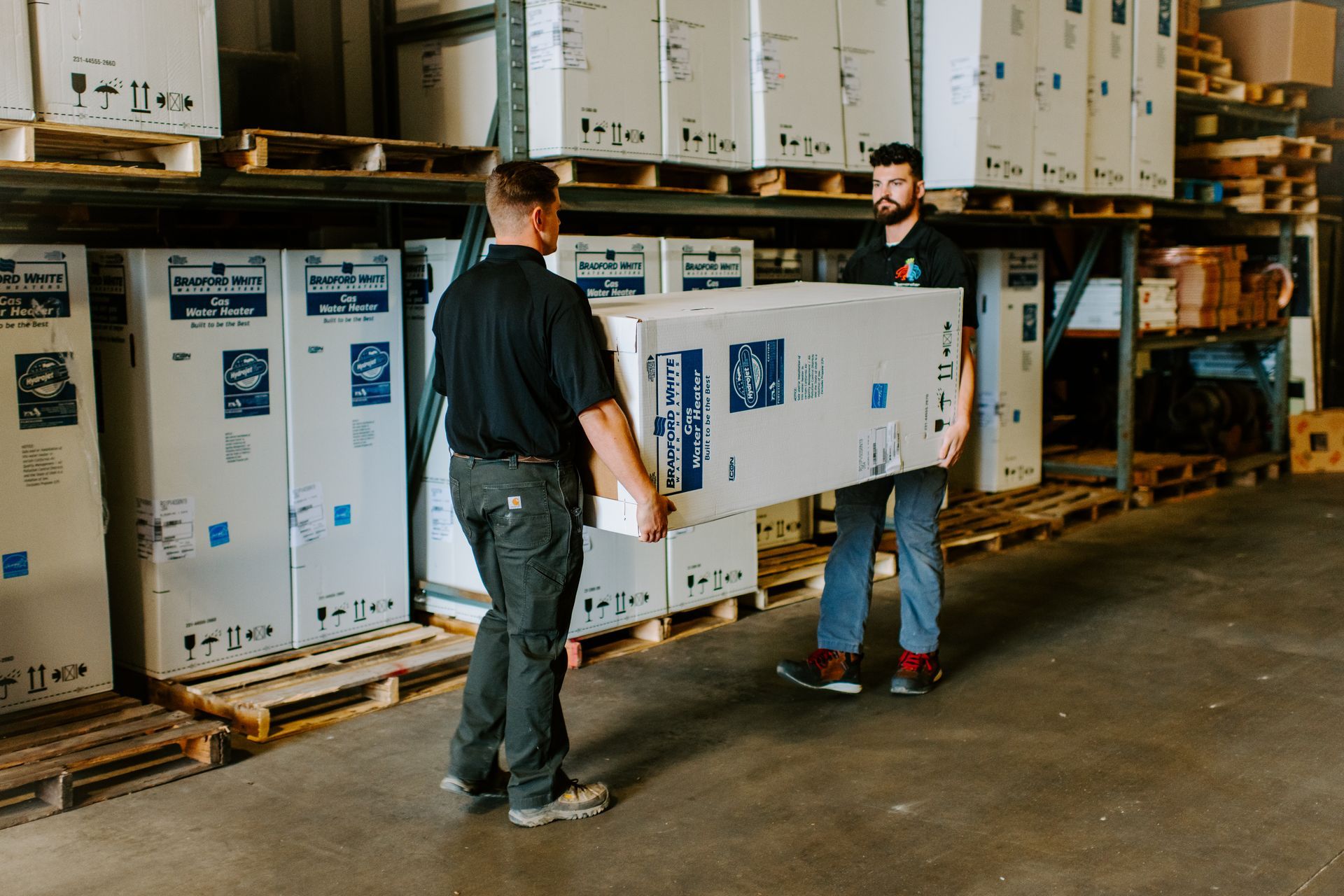 Two men in a warehouse lifting a large white box from a shelf stacked with similar boxes.