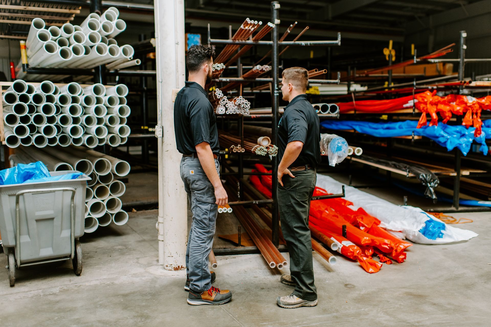 Two men in work clothes discussing pipes in a warehouse setting.