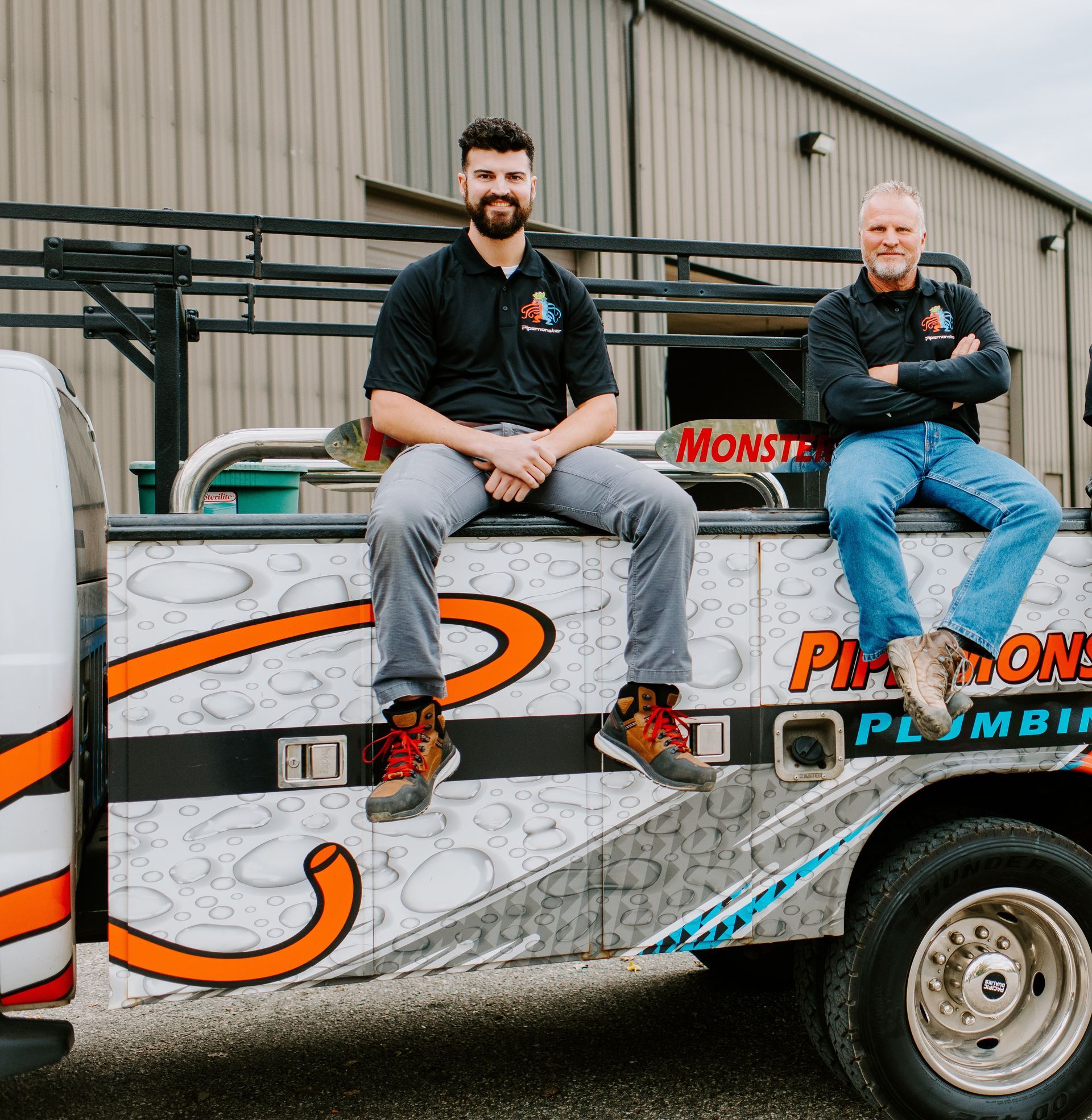 Two men sit on a plumbing truck bed, smiling. One younger, in gray pants; the other older, in jeans.
