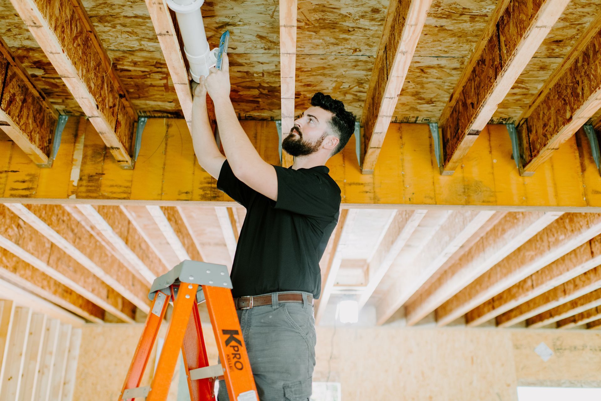 A man is standing on a ladder fixing a pipe on the ceiling.