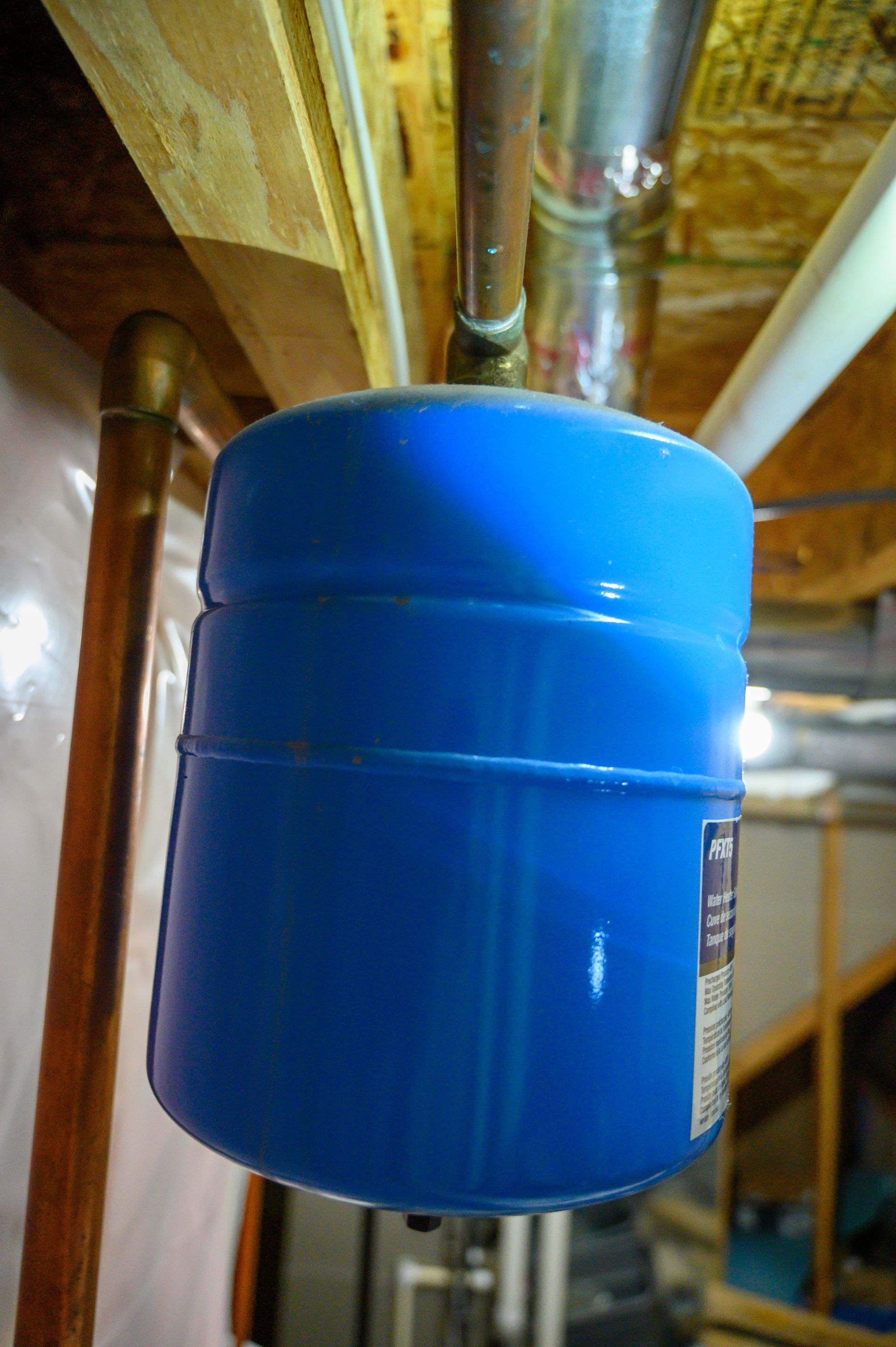 A blue water tank is hanging from a pipe in a basement.