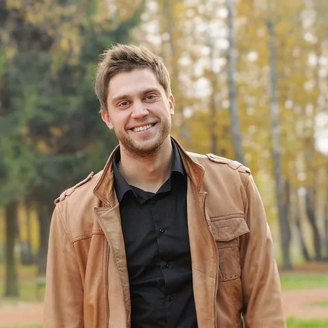 A man wearing a brown jacket and a black shirt is smiling for the camera.