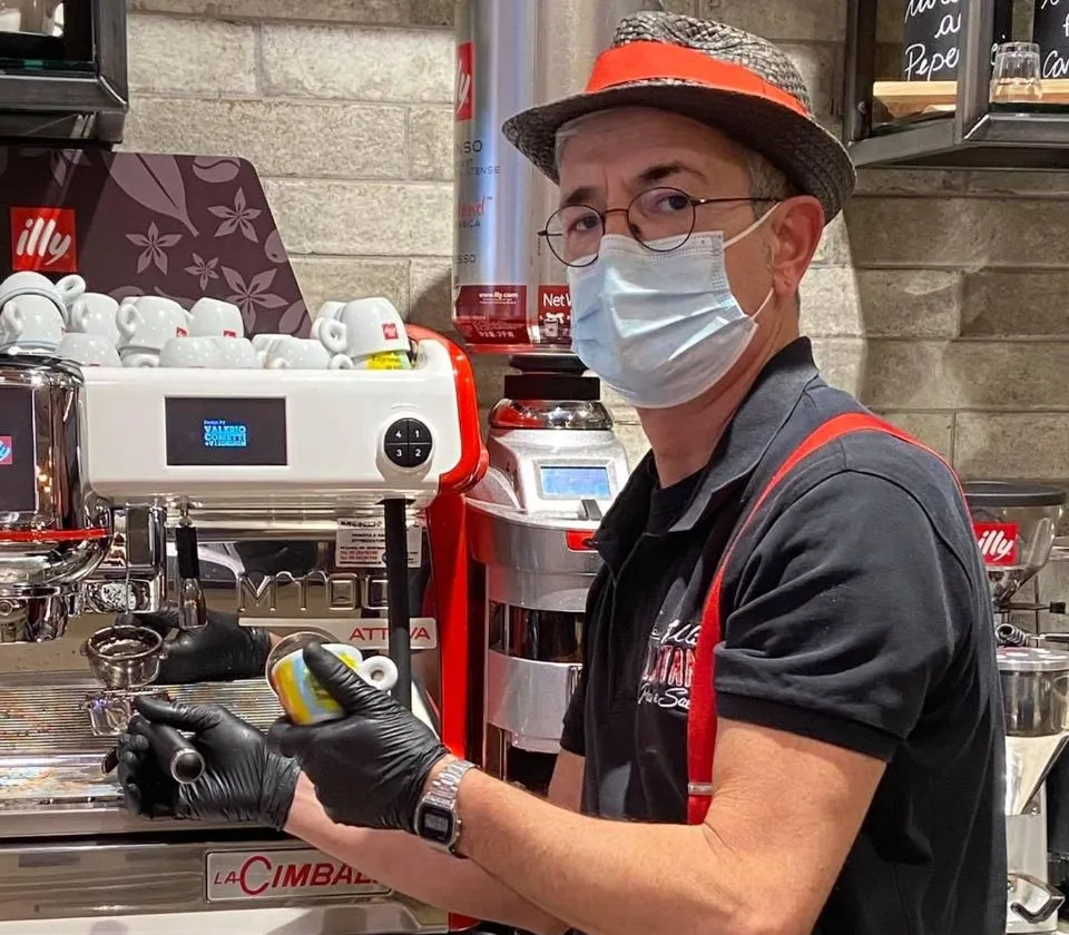 Un uomo che indossa una maschera e un cappello è in piedi davanti a una macchina del caffè.