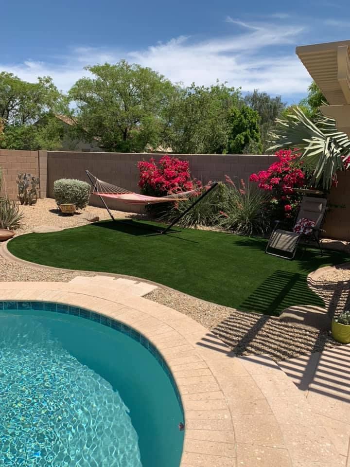 Backyard with pool, green turf, hammock, red flowers, and lounge chair under a blue sky.