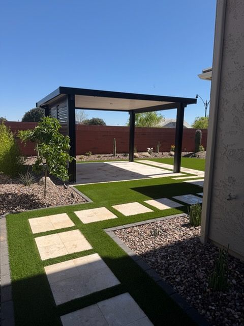 A black modern pergola stands on a patio with pavers and artificial turf in a sunny backyard with a red brick wall.