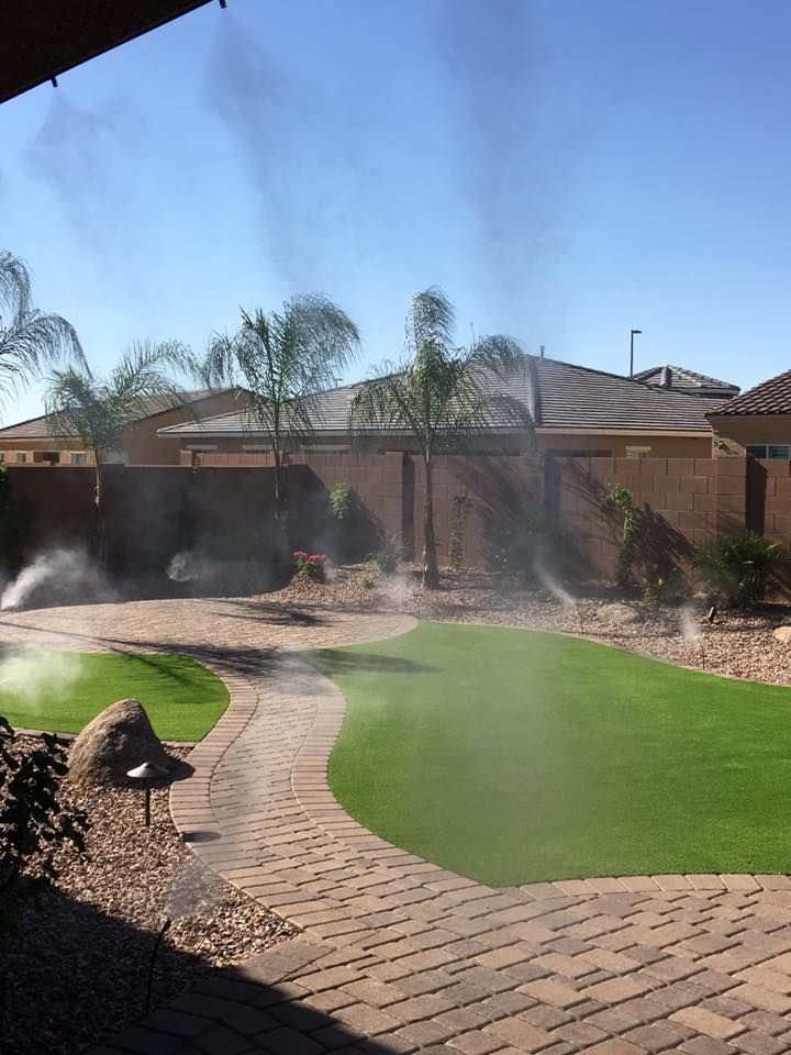 Backyard with misting system, brick patio, green lawn, palm trees, and houses in the background.