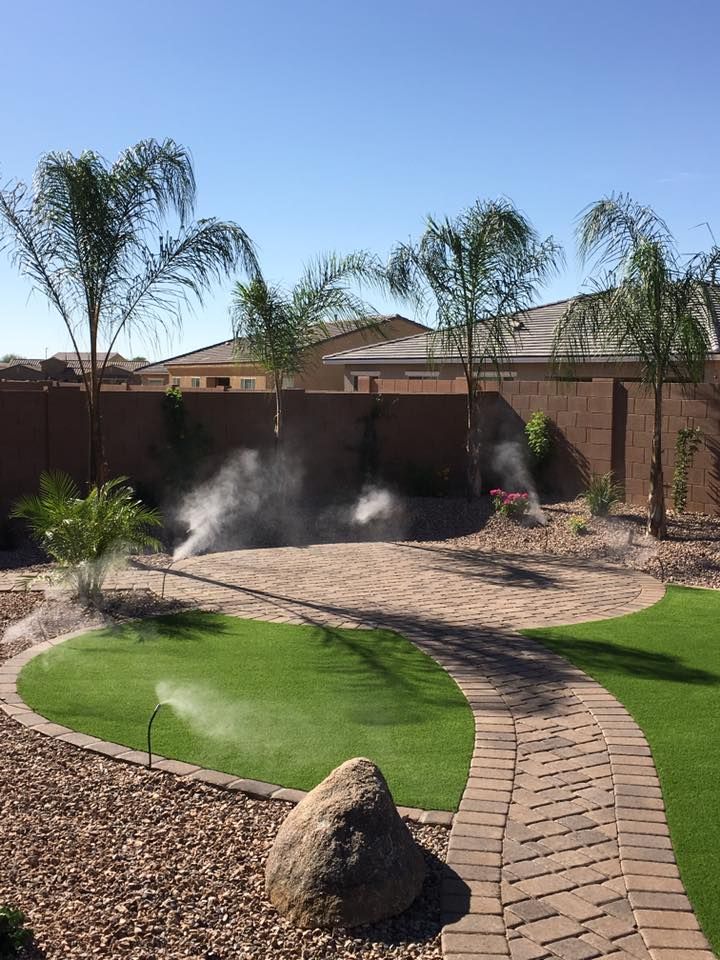 Backyard with sprinklers, artificial grass, and a brick pathway under a sunny blue sky.