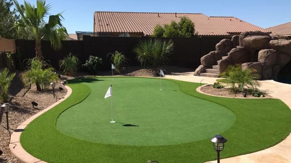 Backyard putting green with two white flags, surrounded by green turf and landscaping.