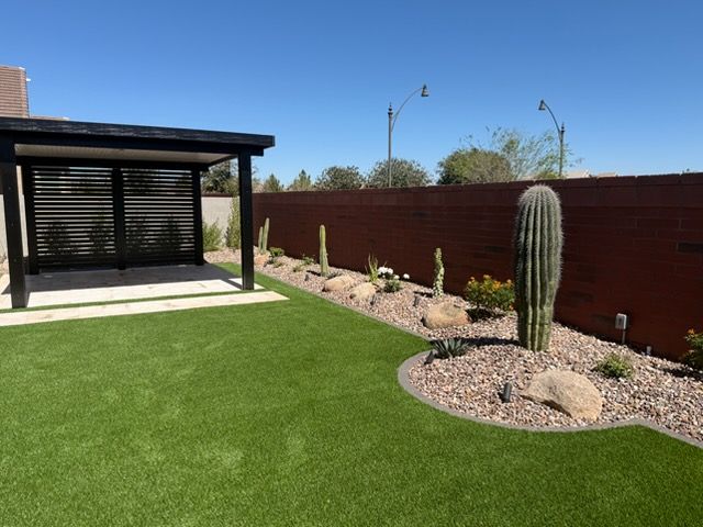 A modern backyard with artificial turf, a black pergola, a brown privacy wall, and desert landscaping with a tall cactus.