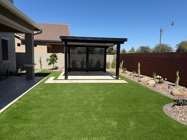 A modern backyard features green artificial turf, a black pergola, a stone patio, and a desert-landscaped border.