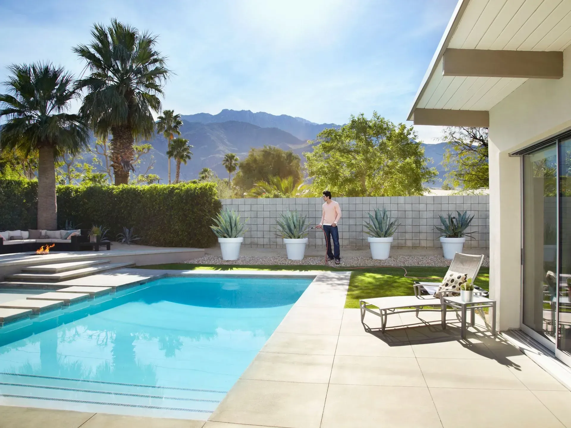 Poolside view: man, modern home, pool, potted plants, palm trees, mountains.