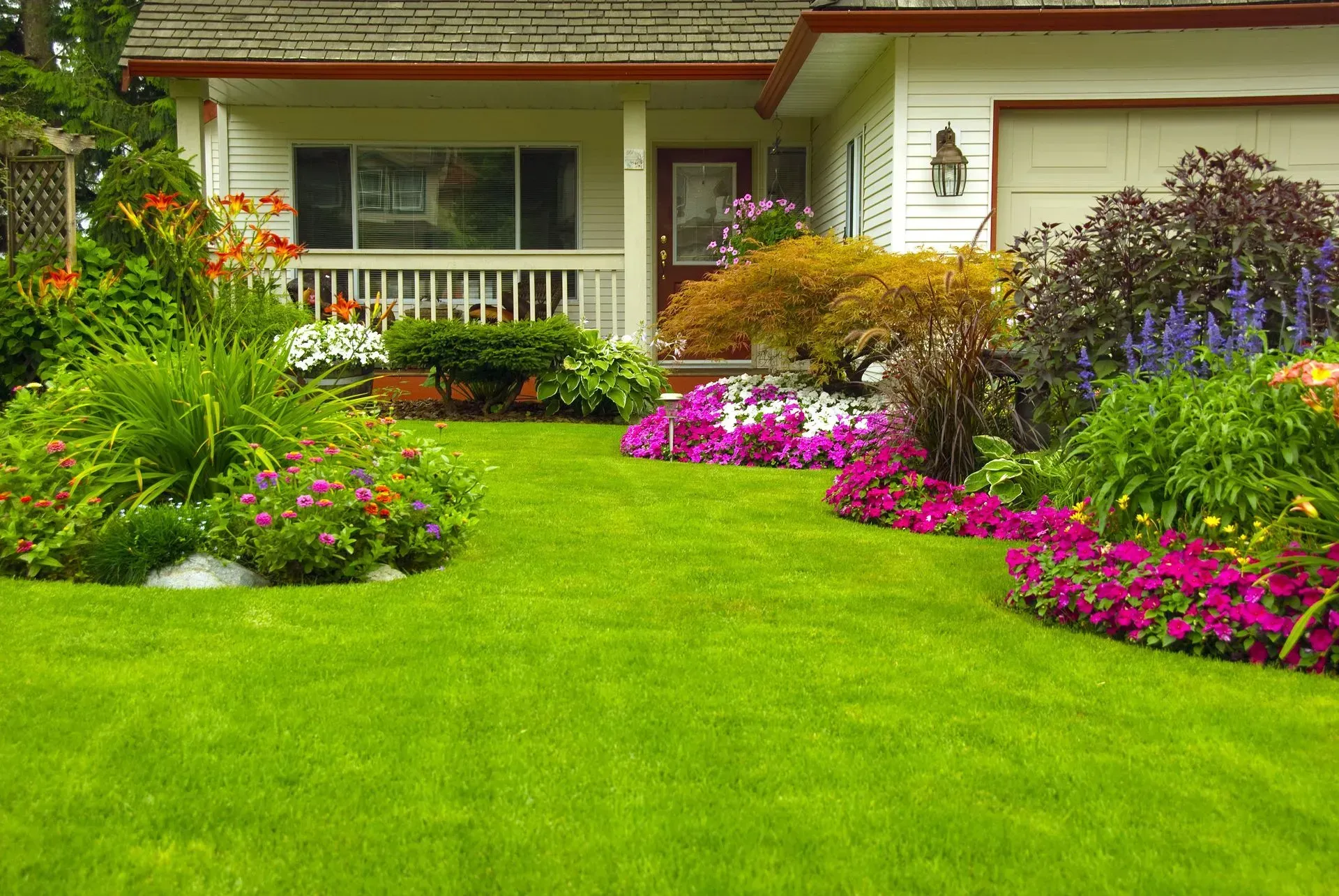 Well-manicured lawn and flowerbeds in front of a white house with a porch and garage.