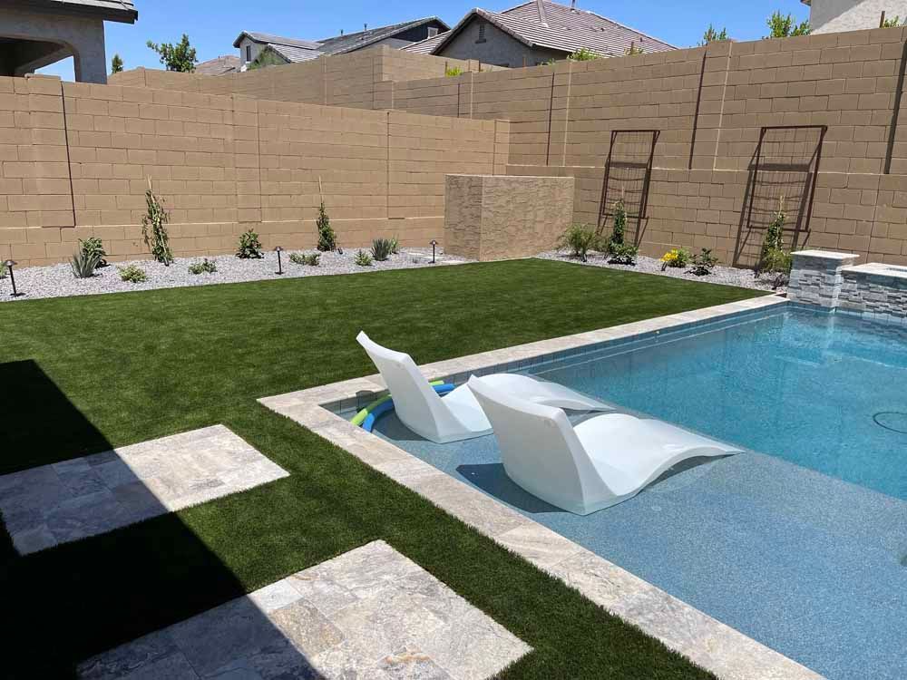 Backyard pool with white lounge chairs, green grass, and a beige block wall.
