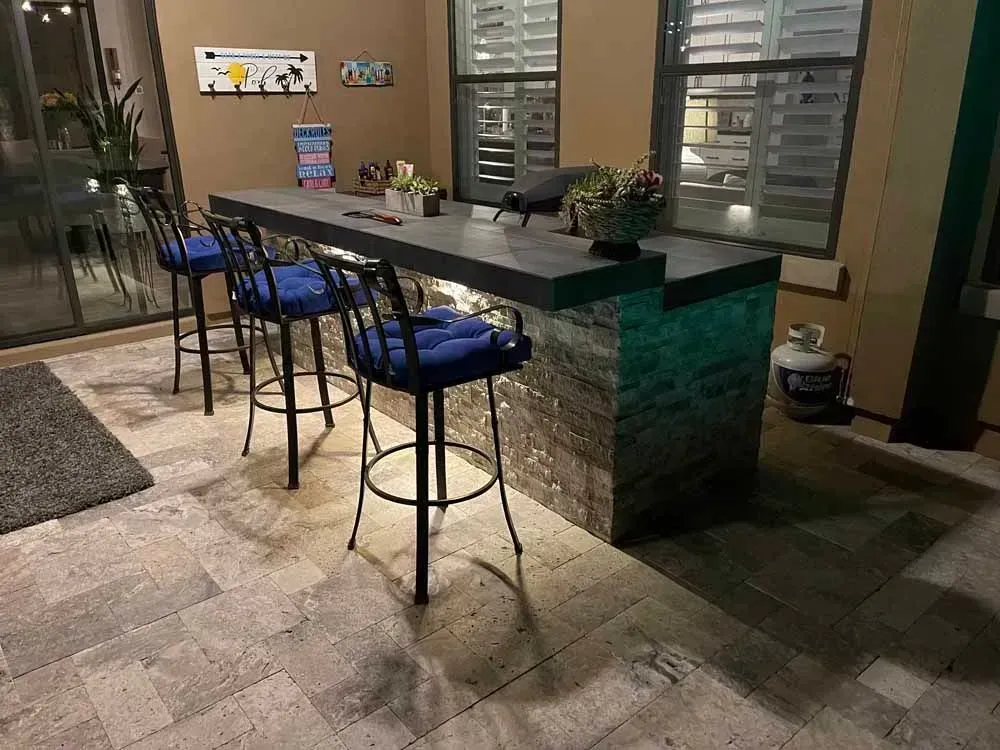 Outdoor bar with stone facade, dark countertop, and black bar stools with blue cushions.