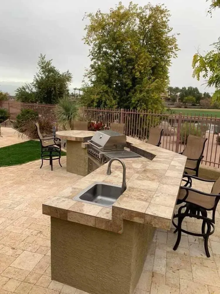 Outdoor kitchen with a grill, sink, and bar seating on a stone patio. Overcast sky.