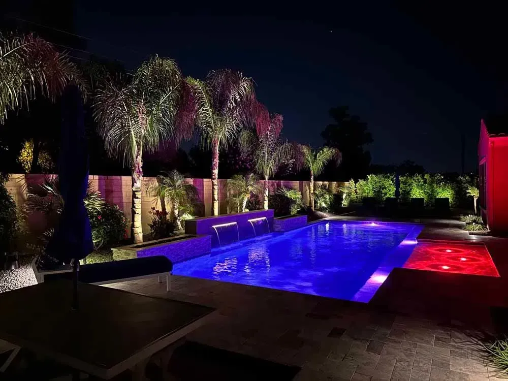 Nighttime view of a blue-lit spool with a waterfall feature, and pink-lit palm trees, surrounded by an illuminated patio.