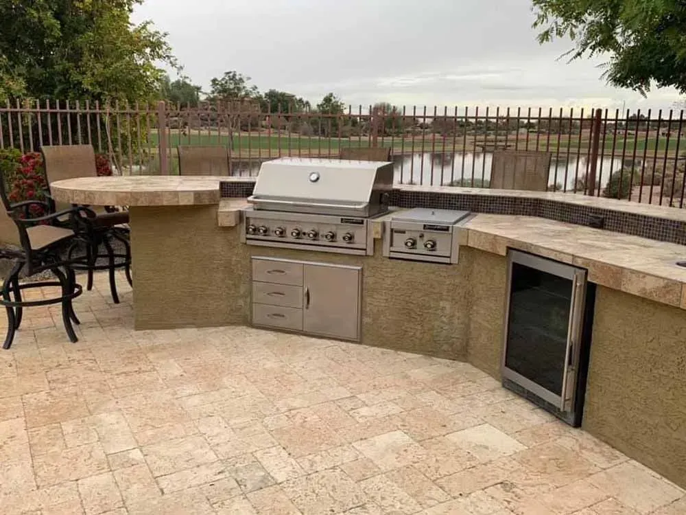 Outdoor kitchen with grill, fridge, and countertop by a lake.