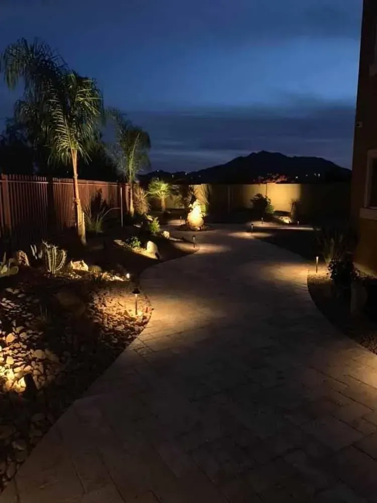 A nighttime view of a lit pathway leading through a yard with landscaping and a mountain in the distance.