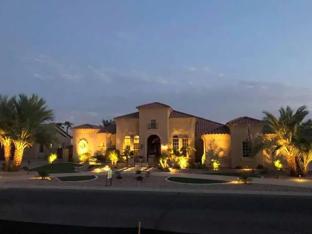 House exterior at dusk, illuminated by warm outdoor lights. Palm trees flank the building.