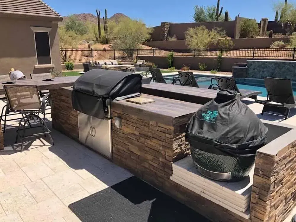 Outdoor kitchen with grills and seating near a pool in a desert landscape.