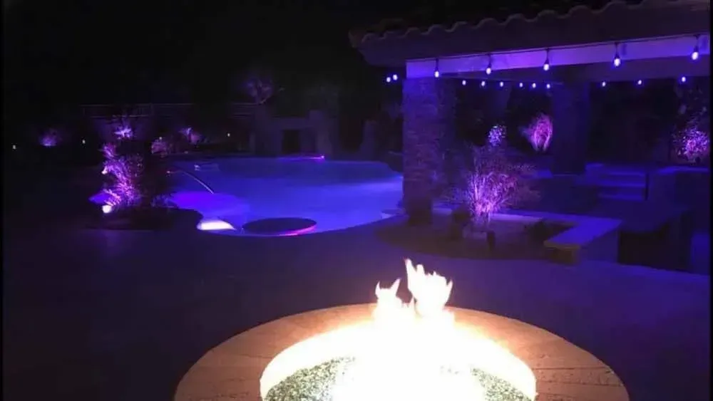 Night scene with a lit fire pit in the foreground and a purple-lit pool and patio area in the background.