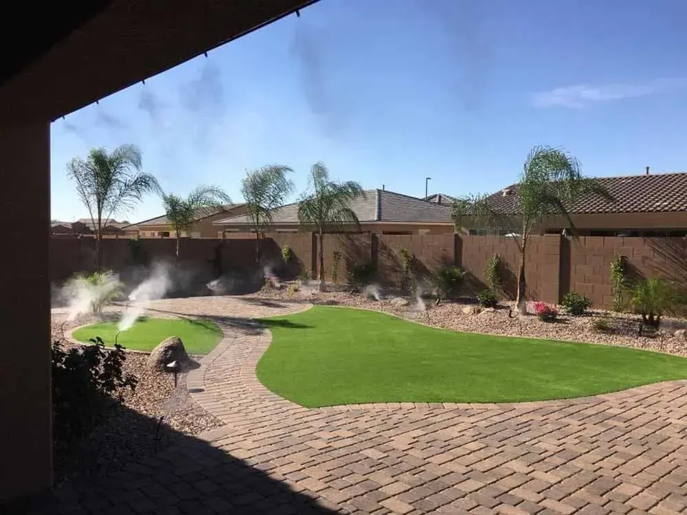 Backyard with misting system, green lawn, palm trees, and brick patio under a sunny sky.