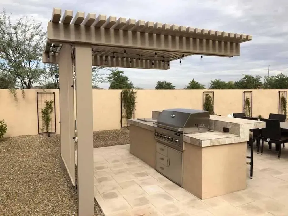 Outdoor kitchen under a beige pergola with built-in grill, stone countertop, and tan cabinetry.