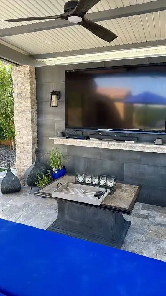 Patio with large TV, stone wall, fan, and blue outdoor furniture.