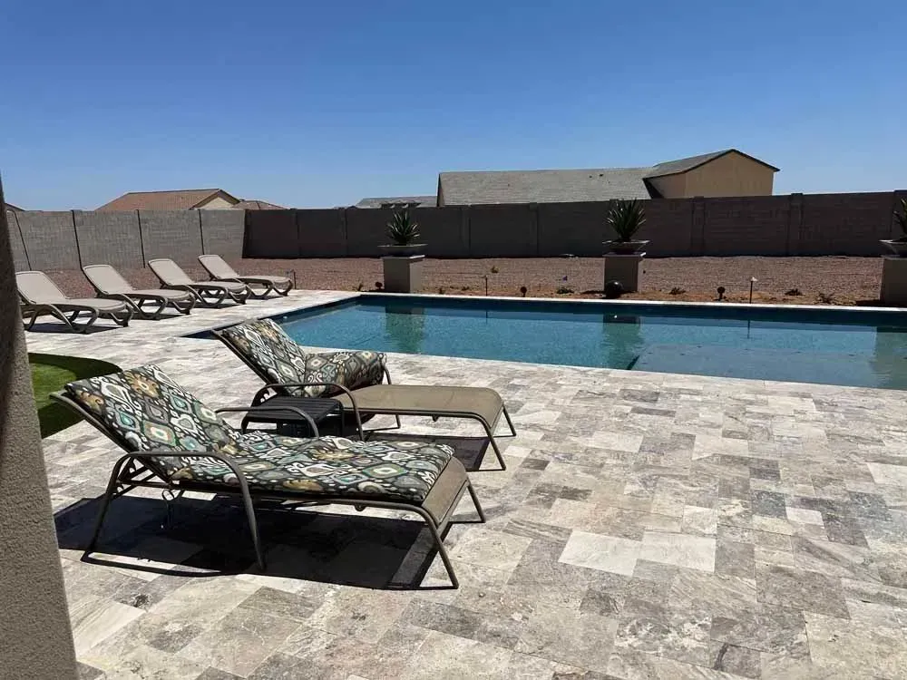 Swimming pool with lounge chairs on a stone patio, surrounded by a wall and clear blue sky.
