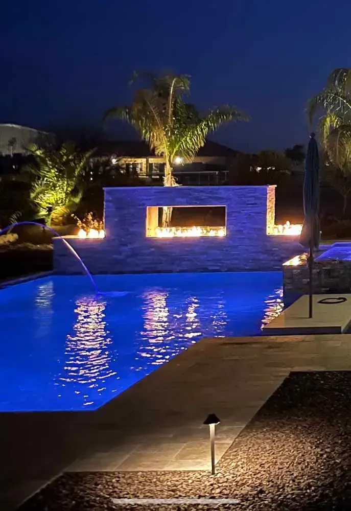 Pool lit up at night with fire features; blue water, stone wall, palm trees.