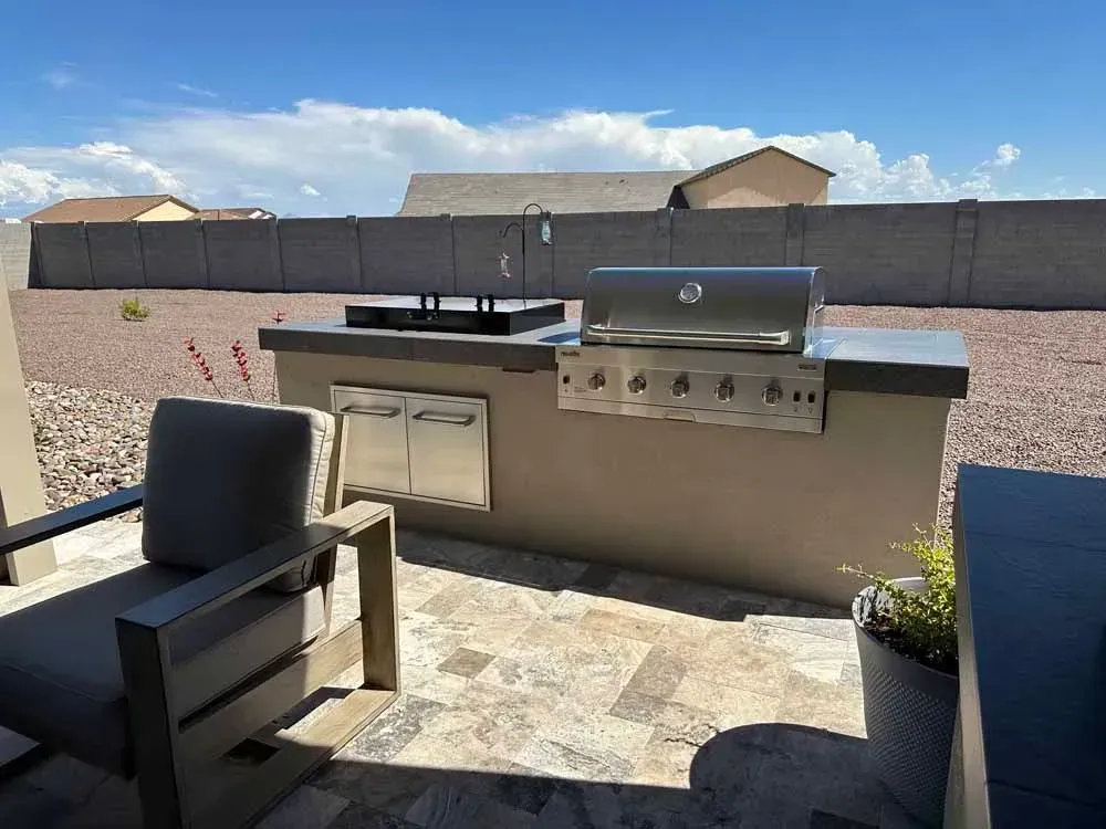 Outdoor kitchen with a grill, sink, and storage, on a stone patio with a chair.