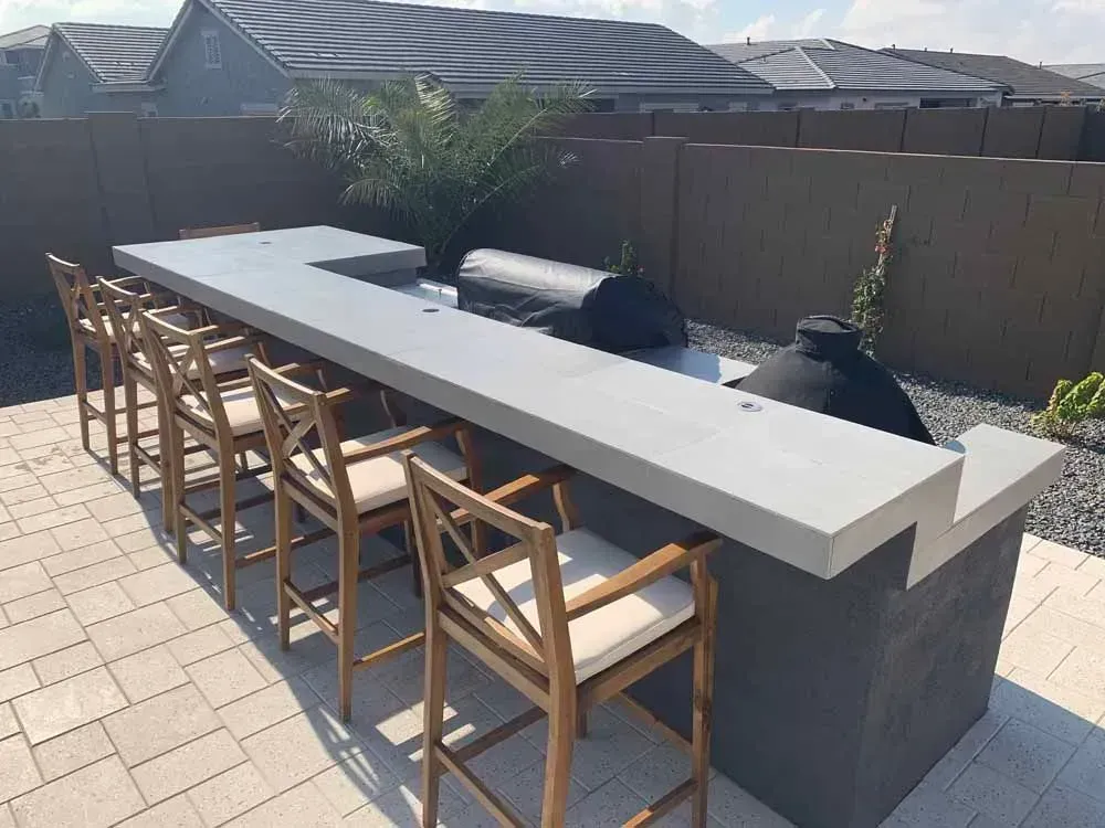 Outdoor bar with concrete countertop, seating, and grill; against a fence.