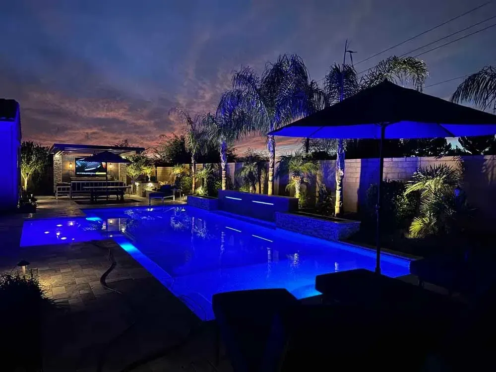Nighttime view of a blue-lit pool surrounded by landscaping, an outdoor kitchen, and patio furniture.