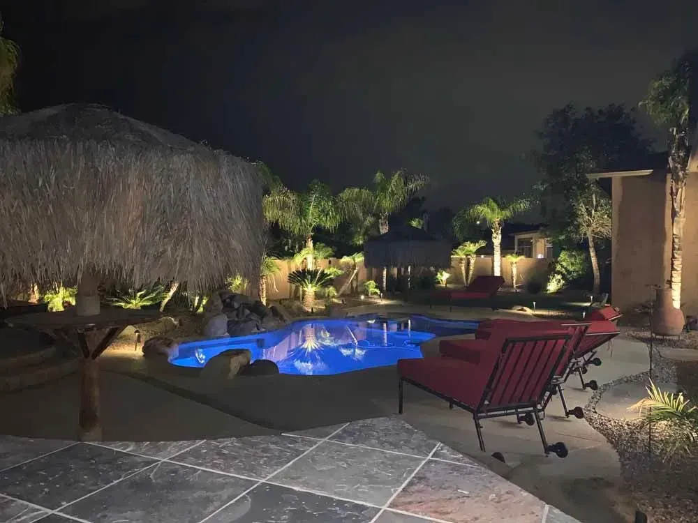 Night view of a backyard pool with blue lights, tiki hut, red lounge chairs, and landscape lighting.