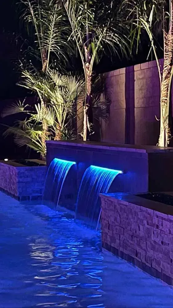 Nighttime shot of a pool with glowing blue waterfalls and palm trees, lit with purple lights against a dark background.