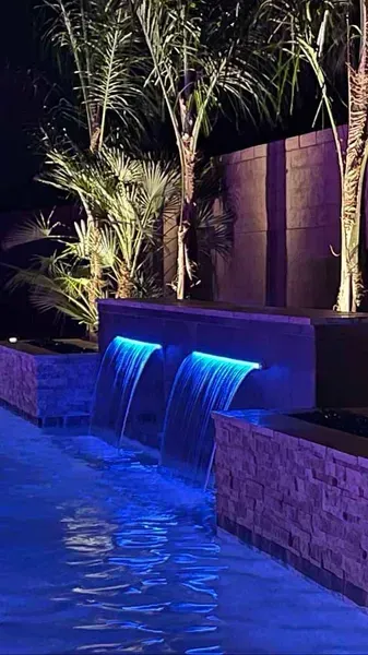 Blue-lit waterfalls cascade into a pool at night, with palm trees and a stone wall illuminated in the background.