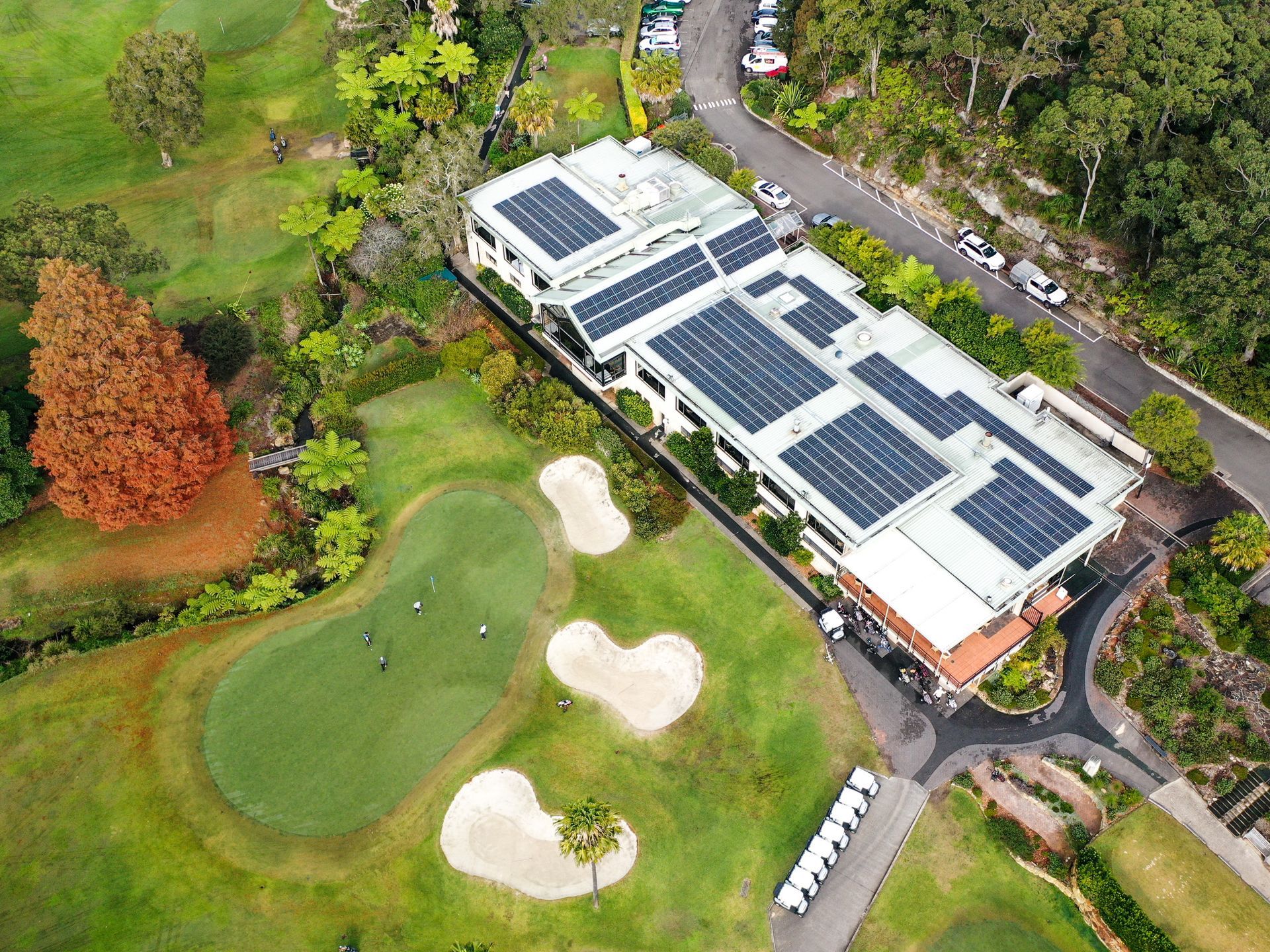 Aerial view of a building with solar panels beside a golf course and winding road.
