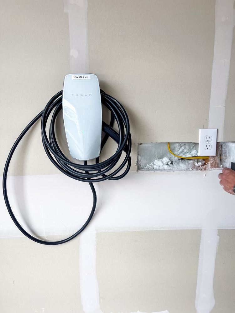 Wall-mounted EV charger with coiled black cable on a garage wall beside an outlet box