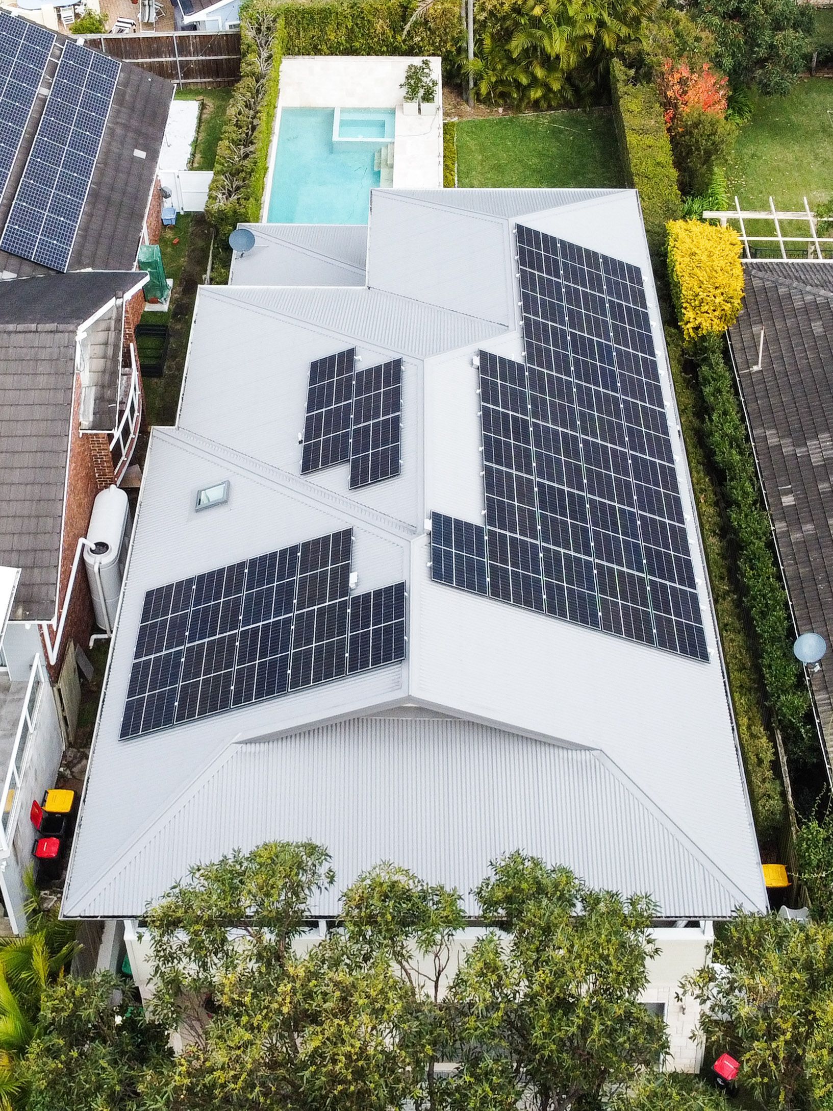 Aerial view of a house with a gray roof covered in solar panels, surrounded by trees and a backyard pool.