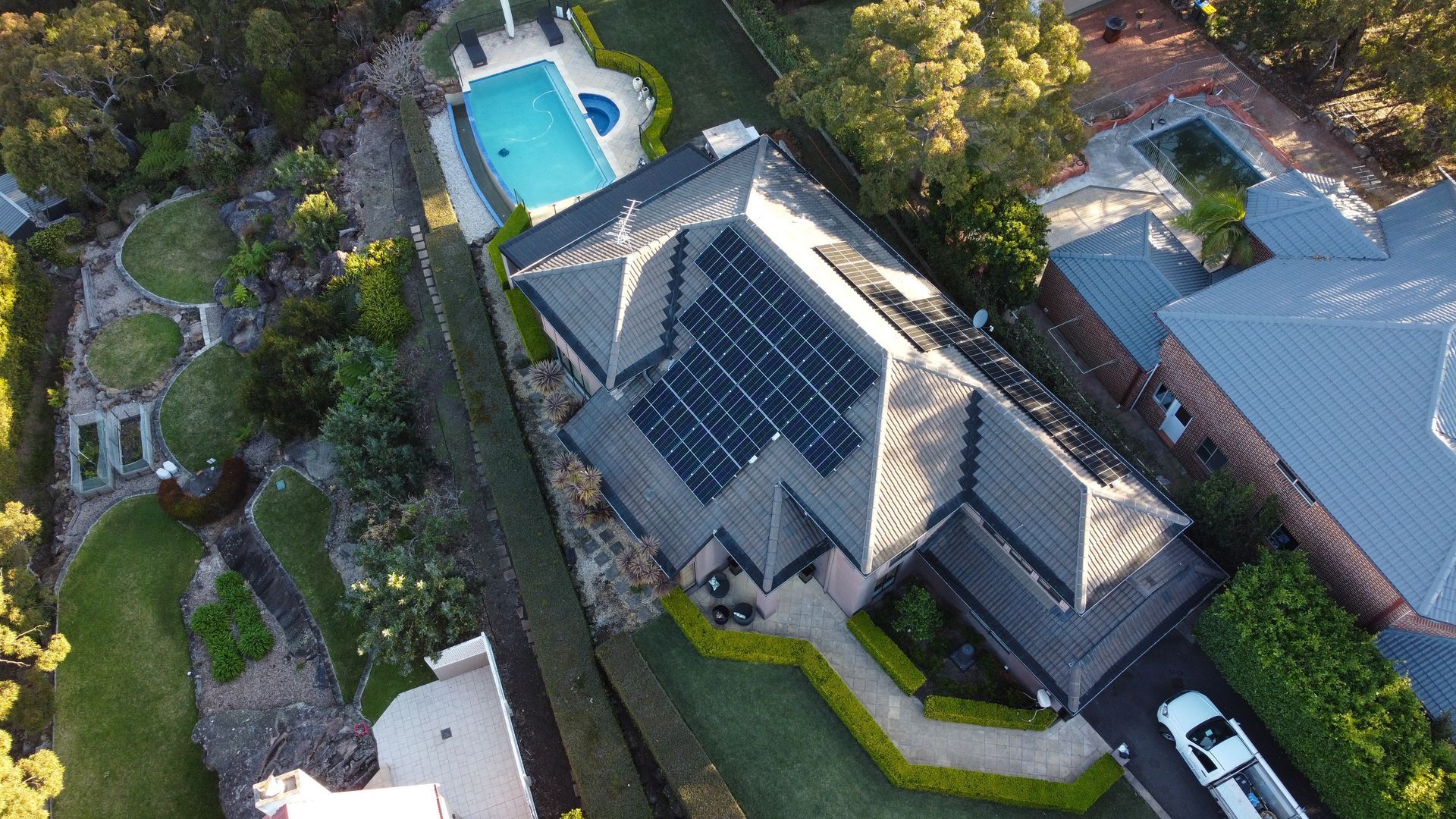Aerial view of a house with solar panels, a pool, and surrounding trees and driveway.