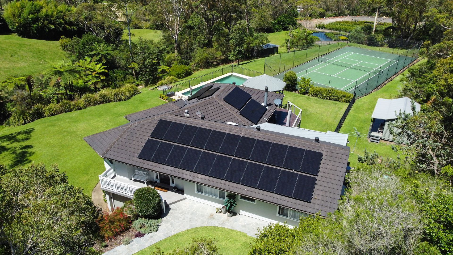 Aerial view of a house with solar panels, surrounded by green lawn and tennis courts